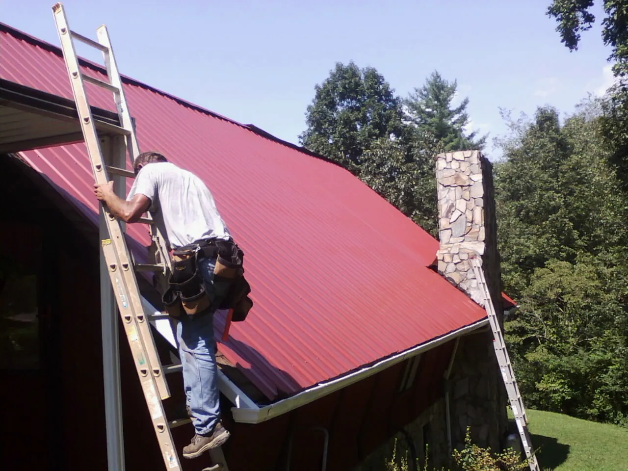 Roofer on a ladder beside a red metal roof and stone chimney — Soffit & Fascia Repair by Roofing Brothers in Schulenburg